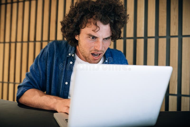 Ritratto di un giovane scioccato dai capelli ricci che lavora sul computer portatile navigando su Internet, leggendo la notizia l immagini stock libere da diritti Ritratto di un giovane scioccato dai capelli ricci che lavora sul computer portatile navigando su Internet, leggendo la notizia l immagini stock libere da diritti