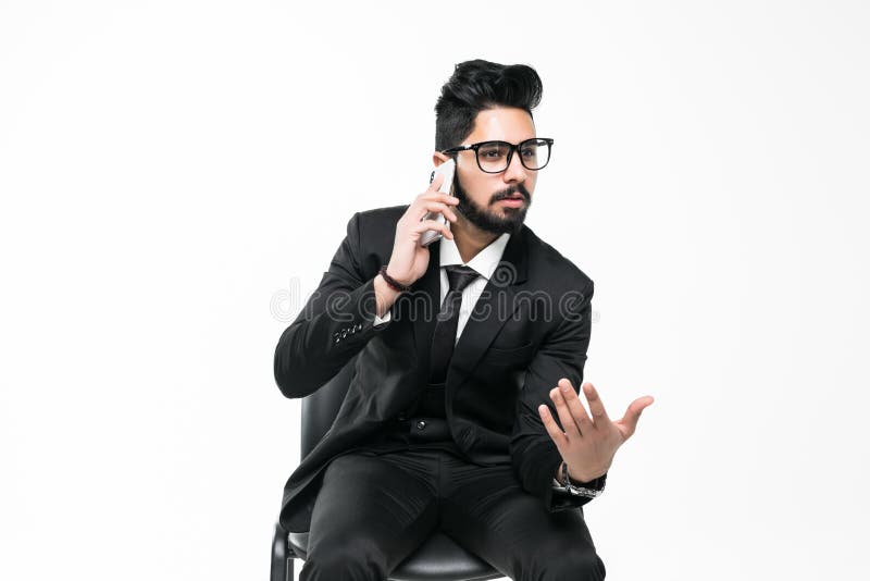 Ritratto di un uomo d'affari arabo o indiano con gli occhiali seduto su una sedia che parla al telefono isolato su fondo bianco fotografia stock libera da diritti Ritratto di un uomo d'affari arabo o indiano con gli occhiali seduto su una sedia che parla al telefono isolato su fondo bianco fotografia stock libera da diritti