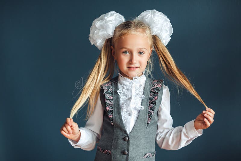 Ritratto di una bambina allegra con gli archi bianchi, la blusa bianca ed il cuoio grigio su fondo blu Sguardo della scolara del fotografie stock libere da diritti Ritratto di una bambina allegra con gli archi bianchi, la blusa bianca ed il cuoio grigio su fondo blu Sguardo della scolara del fotografie stock libere da diritti