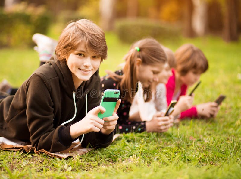 Ritratto di una bambina sorridente che usa il cellulare e si stende con gli amici sull'erba al parco fotografie stock