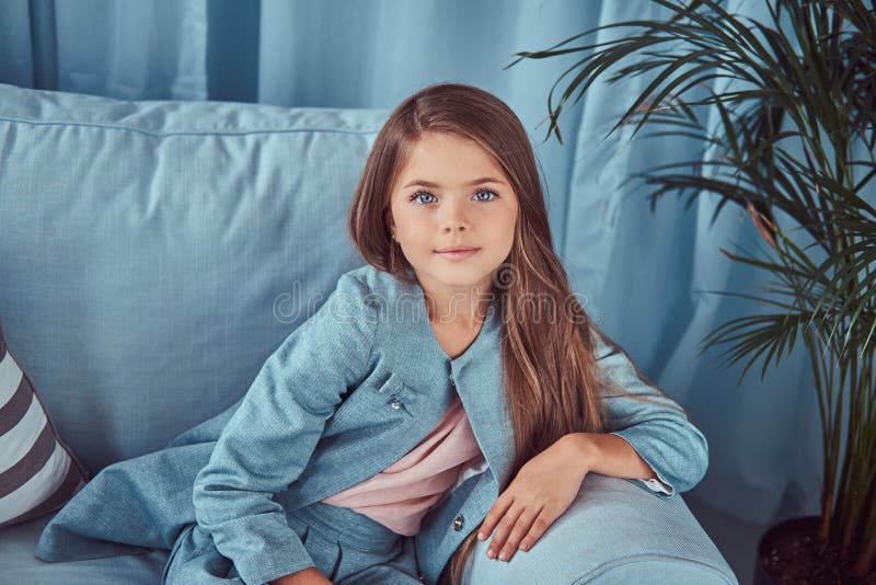 Ritratto di una bambina sveglia che porta un vestito alla moda, sedendosi su un sofà a casa, esaminando una macchina fotografica immagine stock Ritratto di una bambina sveglia che porta un vestito alla moda, sedendosi su un sofà a casa, esaminando una macchina fotografica immagine stock