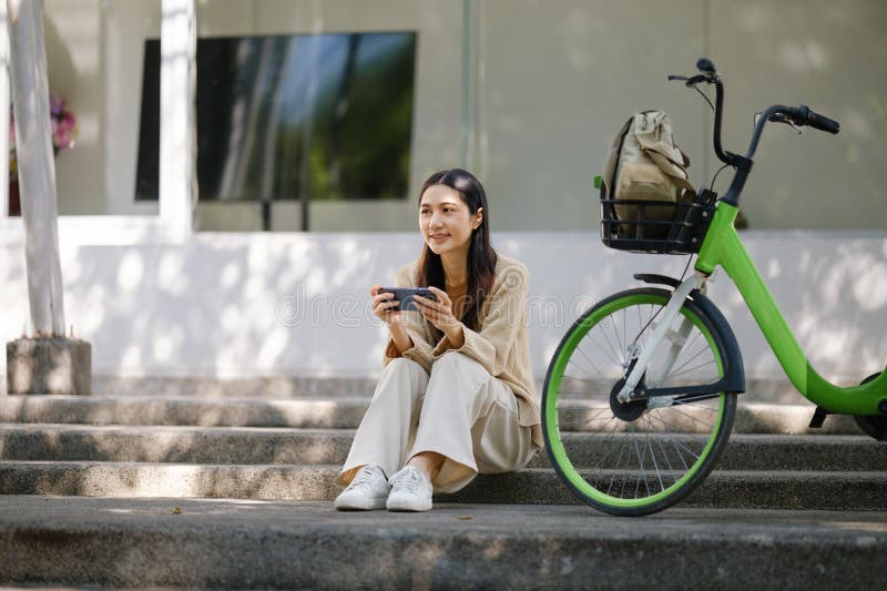Ritratto di una donna asiatica allegra che gioca sui social media dallo smartphone e siediti vicino alla bicicletta per terra immagini stock libere da diritti