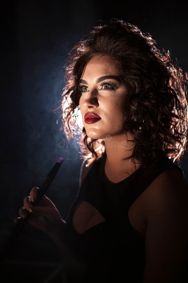 Ritratto di una donna in una barra del narghilé Donna che porta il vestito profondo dalla scollatura Trucco luminoso e acconciatu fotografie stock Ritratto di una donna in una barra del narghilé Donna che porta il vestito profondo dalla scollatura Trucco luminoso e acconciatu fotografie stock