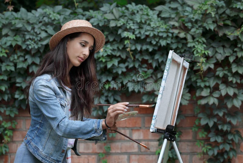 Ritratto di una giovane donna carina in quel quadro che dipinge con pennelli e un moderno cavalletto sulla natura in un parco esti immagini stock Ritratto di una giovane donna carina in quel quadro che dipinge con pennelli e un moderno cavalletto sulla natura in un parco esti immagini stock