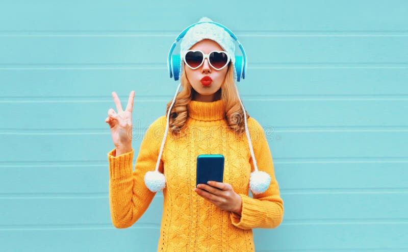 Winter portrait cool woman with phone in headphones listening to music showing peace hand sign blowing red lips wearing yellow knitted sweater, white hat, heart shaped sunglasses on blue background. Winter portrait cool woman with phone in headphones listening to music showing peace hand sign blowing red lips wearing yellow knitted sweater, white hat, heart shaped sunglasses on blue background