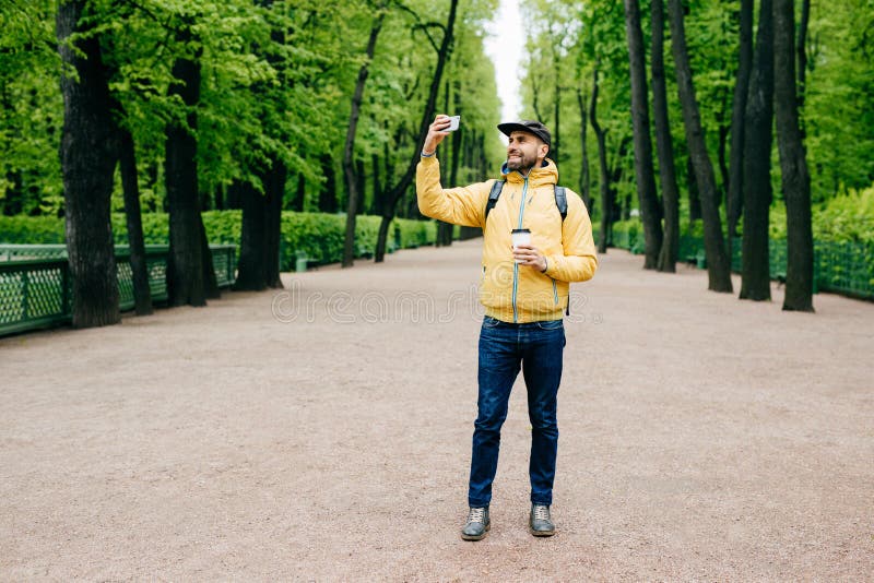Ritratto orizzontale dell'uomo barbuto bello in giacca a vento, cappuccio giallo e jeans aventi espressione felice mentre posando fotografia stock Ritratto orizzontale dell'uomo barbuto bello in giacca a vento, cappuccio giallo e jeans aventi espressione felice mentre posando fotografia stock