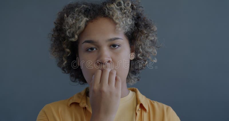 Ritratto in slow motion di una giovane donna africana nervosa, afro-americana che morde le unghie sentendo ansia sullo sfondo grig