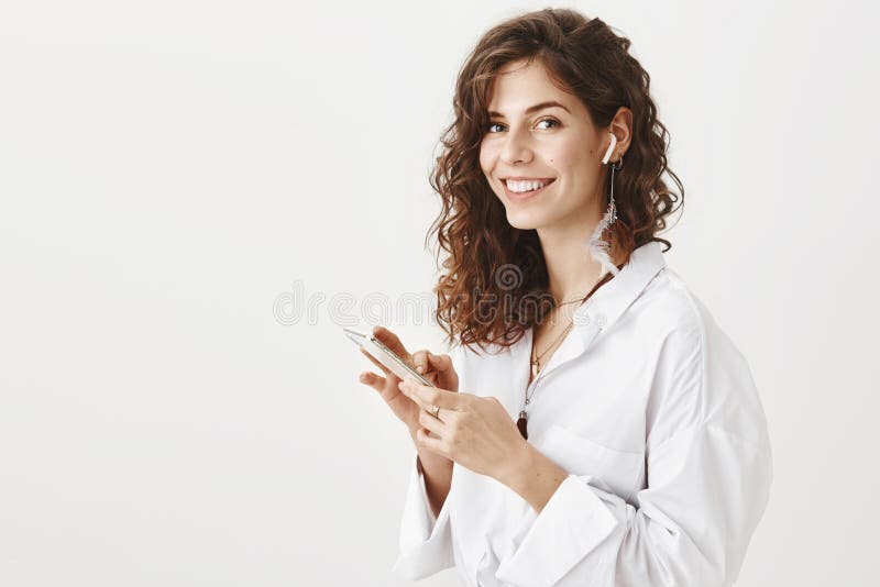 Riuscita donna caucsian positiva che parla sullo smartphone via i earbuds senza fili, dispositivo di tenuta a disposizione mentre fotografia stock libera da diritti Riuscita donna caucsian positiva che parla sullo smartphone via i earbuds senza fili, dispositivo di tenuta a disposizione mentre fotografia stock libera da diritti