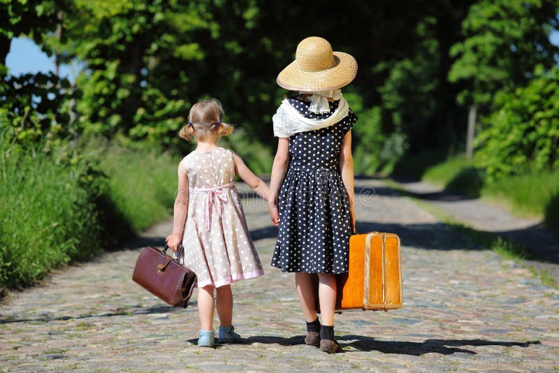 Due bambine, camminando con il suo bagaglio in un viale alberato, visto dal retro.