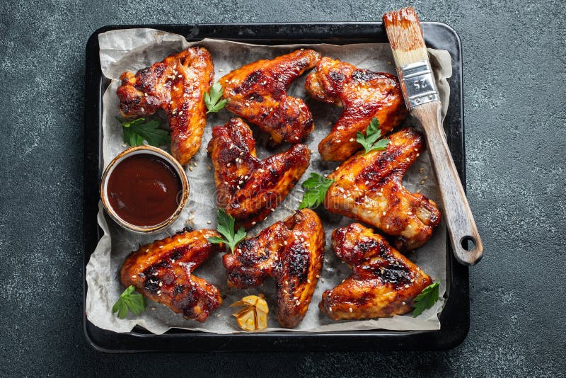 Roasted chicken wings in barbecue sauce with sesame seeds and parsley in a baking tray on a dark table. Top view. Tasty snack for