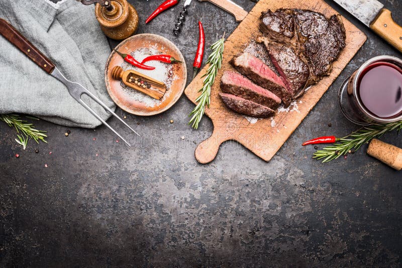 Roasted sliced grill steak on wooden cutting board with wine, seasoning and meat fork on dark vintage metal background, top view