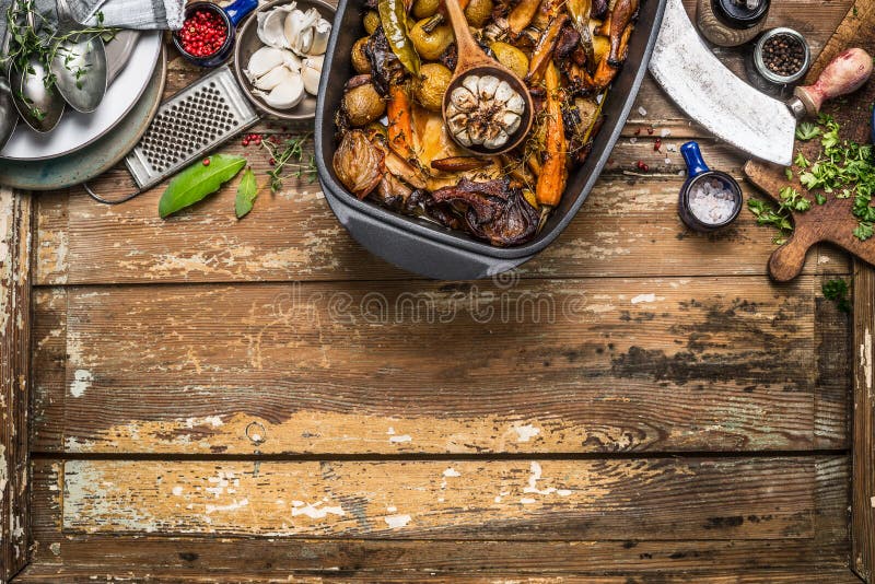 Roasted vegetables stew in cooking pot with kitchen tools on rustic wooden background, top view