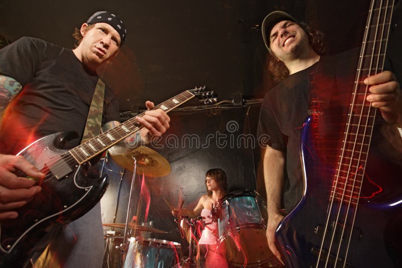 Band playing on a stage. Guitarist, bassist and female drummer. Shot with strobes and slow shutter speed to create lighting atmosphere and blur effects. Slight motion blur on performers. Band playing on a stage. Guitarist, bassist and female drummer. Shot with strobes and slow shutter speed to create lighting atmosphere and blur effects. Slight motion blur on performers.