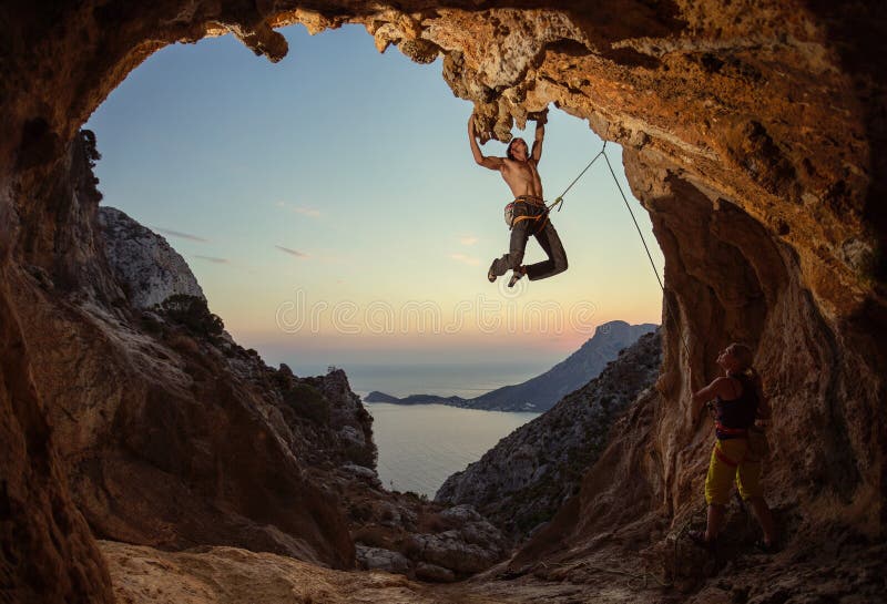 Rock climbing at sunset. Young man climbing route in cave, female partner belaying him