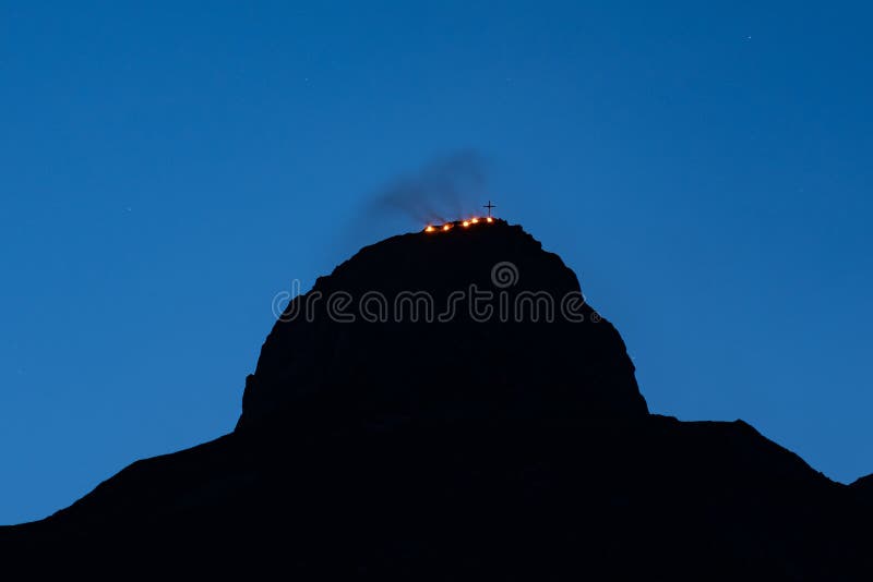 Rock silhouette with bonfires and a cross on top of it under a blue sky during the evening