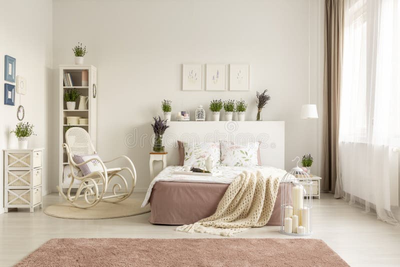 Rocking chair next to bed with blanket in spacious white bedroom interior with pink carpet. Real photo concept