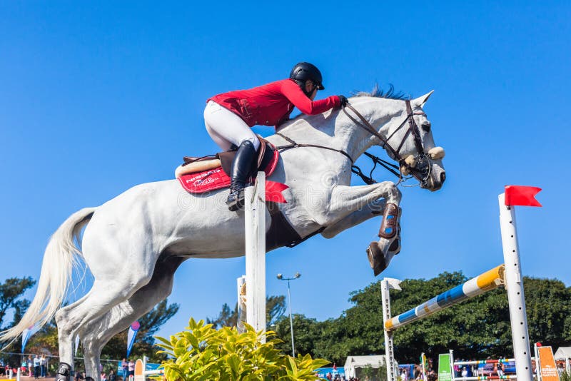 South African Equestrian horse jumping championships held in Durban South-Africa.Close-Up photo of competing female woman rider in red jacket and white horse jumping over poles in mid air flight over gates poles. South African Equestrian horse jumping championships held in Durban South-Africa.Close-Up photo of competing female woman rider in red jacket and white horse jumping over poles in mid air flight over gates poles.