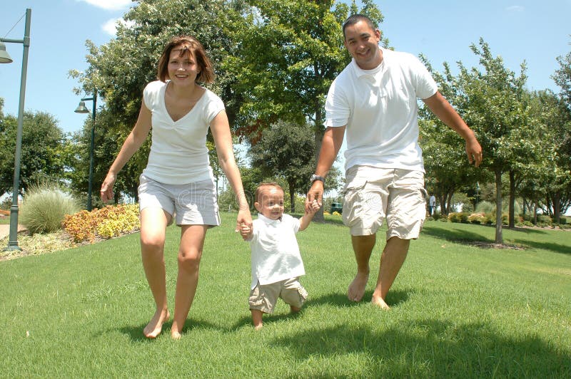 Young married couple with their toddler child. New family taking pictures in white shirts and tan shorts. Family of three holding hands as they walk the park. Young married couple with their toddler child. New family taking pictures in white shirts and tan shorts. Family of three holding hands as they walk the park.