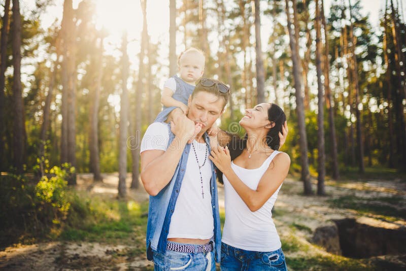 Rodzinna mama, tata i córka, siedzimy przy ojczulkiem na ramionach, i rodzice całują na naturze w lesie w lecie przy słońcem zdjęcia stock Rodzinna mama, tata i córka, siedzimy przy ojczulkiem na ramionach, i rodzice całują na naturze w lesie w lecie przy słońcem zdjęcia stock
