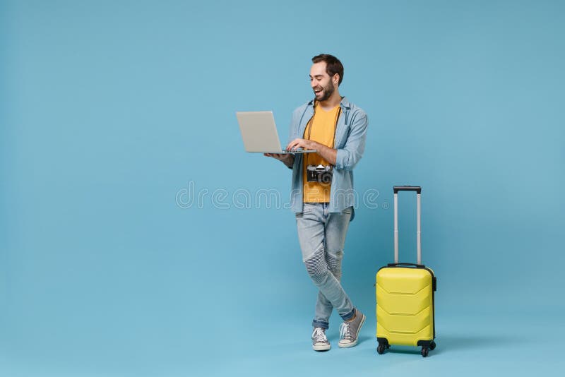 Cheerful traveler tourist man in yellow clothes with photo camera suitcase isolated on blue background. Male passenger traveling abroad on weekend. Air flight journey. Working on laptop booking hotel. Cheerful traveler tourist man in yellow clothes with photo camera suitcase isolated on blue background. Male passenger traveling abroad on weekend. Air flight journey. Working on laptop booking hotel