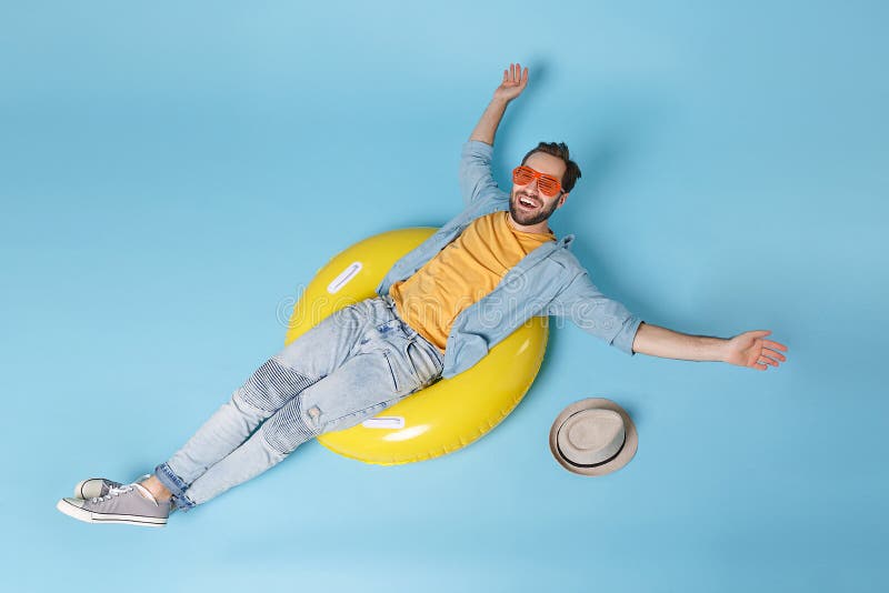 Cheerful traveler tourist man in yellow summer clothes, eyeglasses isolated on blue background. Passenger traveling abroad on weekend. Air flight journey concept. Lying in rubber ring spreading hands. Cheerful traveler tourist man in yellow summer clothes, eyeglasses isolated on blue background. Passenger traveling abroad on weekend. Air flight journey concept. Lying in rubber ring spreading hands