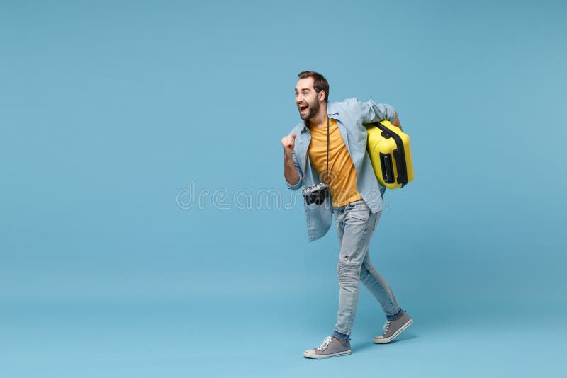 Cheerful traveler tourist man in yellow summer clothes with photo camera isolated on blue background. Male passenger traveling abroad on weekends. Air flight journey concept. Holding suitcase running. Cheerful traveler tourist man in yellow summer clothes with photo camera isolated on blue background. Male passenger traveling abroad on weekends. Air flight journey concept. Holding suitcase running