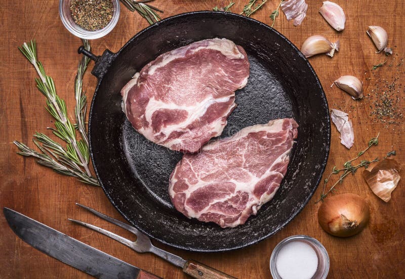 2 rohe frische Schweinefleischsteaks in der alten Gusseisenbratpfanne mit Zwiebelknoblauch und -kräutern auf rustikalem hölzernem lizenzfreie stockfotos 2 rohe frische Schweinefleischsteaks in der alten Gusseisenbratpfanne mit Zwiebelknoblauch und -kräutern auf rustikalem hölzernem lizenzfreie stockfotos