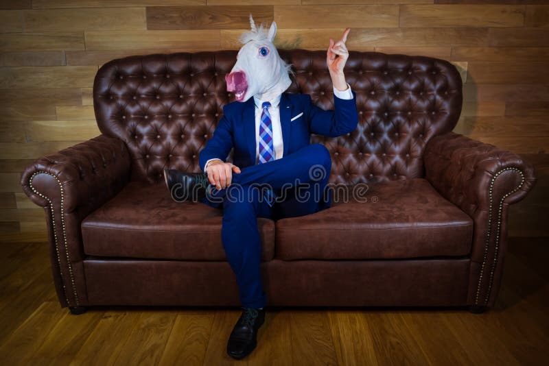 Funny unicorn in elegant suit siting on sofa like a boss and showing hand gesture with raised index finger. Unusual man at home. Freaky guy in mask. Funny unicorn in elegant suit siting on sofa like a boss and showing hand gesture with raised index finger. Unusual man at home. Freaky guy in mask.