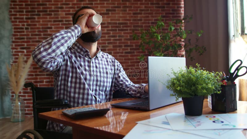 Rollstuhlfahrer arbeiten an einem Laptop, der Kaffee trinkt und nach vorn schaut und lächelt. Hipster-Geschäftsmann nahm Pause ein