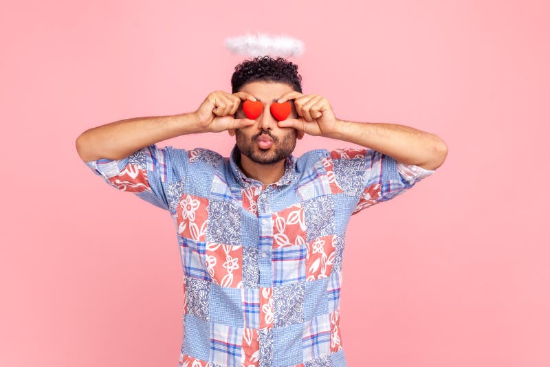 Romantic angelic man with beard covering eyes with small red hearts, making pout lips, kissing