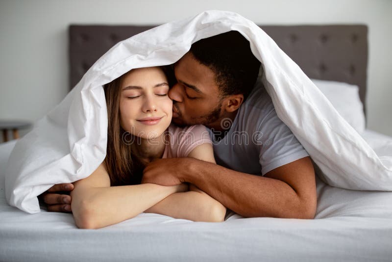 Romantic Black Guy Kissing His Caucasian Girlfriend Under Blanket on Bed, Indoors Stock Image