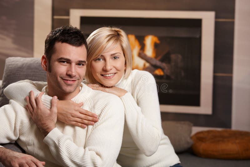 Young couple hugging on sofa in front of fireplace at home, looking at camera, smiling. Young couple hugging on sofa in front of fireplace at home, looking at camera, smiling.