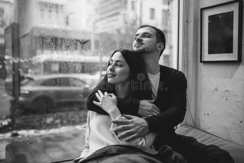 Romantic Couple Loving Guy Hugs His Beautiful Girlfriend Sitting On The Windowsill In A Cozy