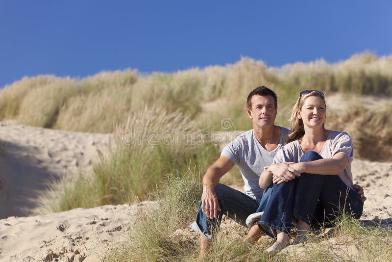 A romantic young man and woman couple sitting together in the sand dunes of a sunny beach with a bright blue sky. A romantic young man and woman couple sitting together in the sand dunes of a sunny beach with a bright blue sky