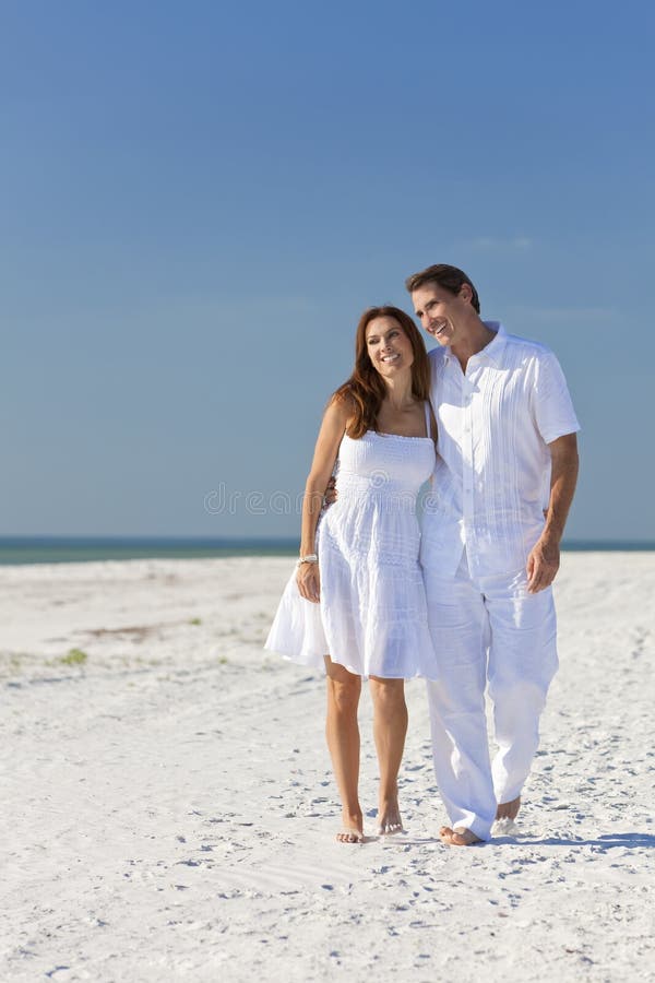 Happy man and woman romantic couple in white clothes walking on a deserted tropical beach with bright clear blue sky. Happy man and woman romantic couple in white clothes walking on a deserted tropical beach with bright clear blue sky