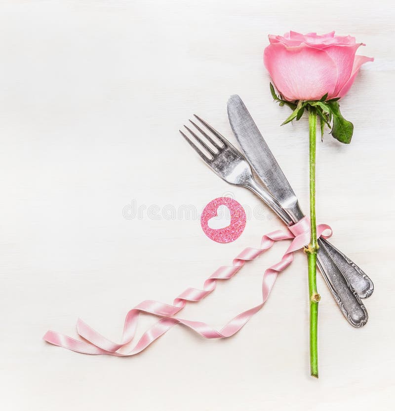 Romantic dinner table place setting with fork, knife , pink rose and heart on white wooden background, top view. Love symbol. Valentine Day or Birthday concept.