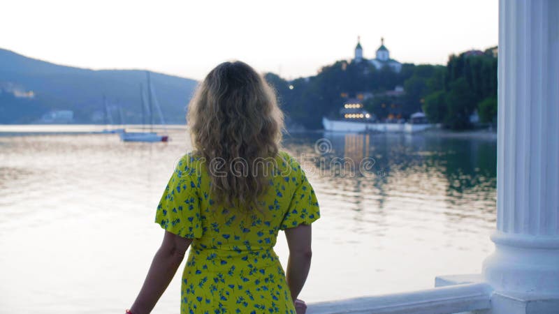 Romantic girl looking to lake and yacht on green hills landscape back view. Relaxing girl admiring evening sea bay on