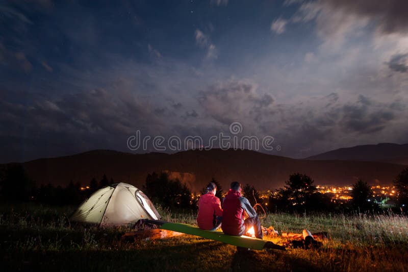 Romantic pair tourists sitting on boards coated camping mat by a bonfire under cloudy sky in the background mountains and luminous town in the evening. Romantic pair tourists sitting on boards coated camping mat by a bonfire under cloudy sky in the background mountains and luminous town in the evening