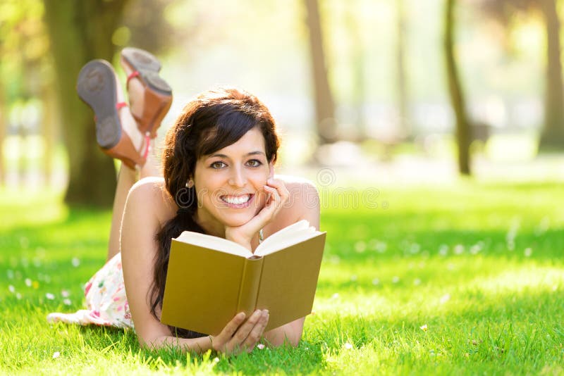 Happy woman reading and holding story book in fresh green park on spring or summer. Caucasian beautifulstudent smiling and day dreaming lying down on grass outdoors in college campus. Happy woman reading and holding story book in fresh green park on spring or summer. Caucasian beautifulstudent smiling and day dreaming lying down on grass outdoors in college campus.