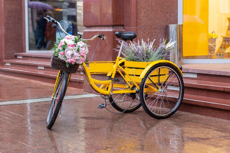 A romantic yellow bike stands on a wet marble pavement after rain. Bicycle with a basket of flowers, lavender and peonies