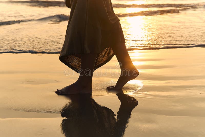 Romantic young girl walks on the beach barefoot in the water. Woman stroll barefoot in the sea at sunset.