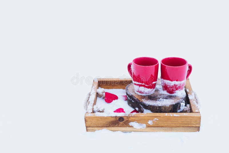 Two red cups with tea on a wooden tray in snow. Romantic Valentine`s day winter picnic, hot tea, couple. Lifestyle, time together outdoors, Valentine`s day. Snowflakes, hoarfrost and heart shape on a tray. Copy space. Two red cups with tea on a wooden tray in snow. Romantic Valentine`s day winter picnic, hot tea, couple. Lifestyle, time together outdoors, Valentine`s day. Snowflakes, hoarfrost and heart shape on a tray. Copy space