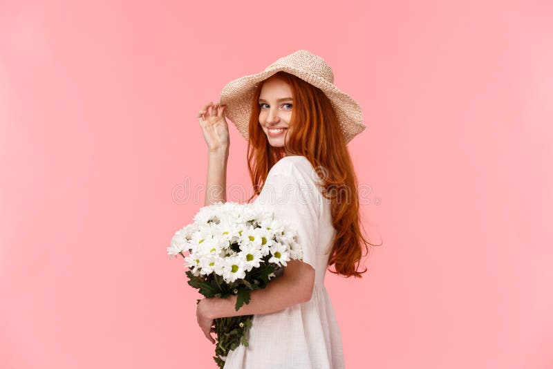 Romantic, silly and tender feminine redhead woman in cute hat, dress, holding bouquet white flowers, turn camera and smiling coquettish, flirting with boyfriend over pink background. Romantic, silly and tender feminine redhead woman in cute hat, dress, holding bouquet white flowers, turn camera and smiling coquettish, flirting with boyfriend over pink background.