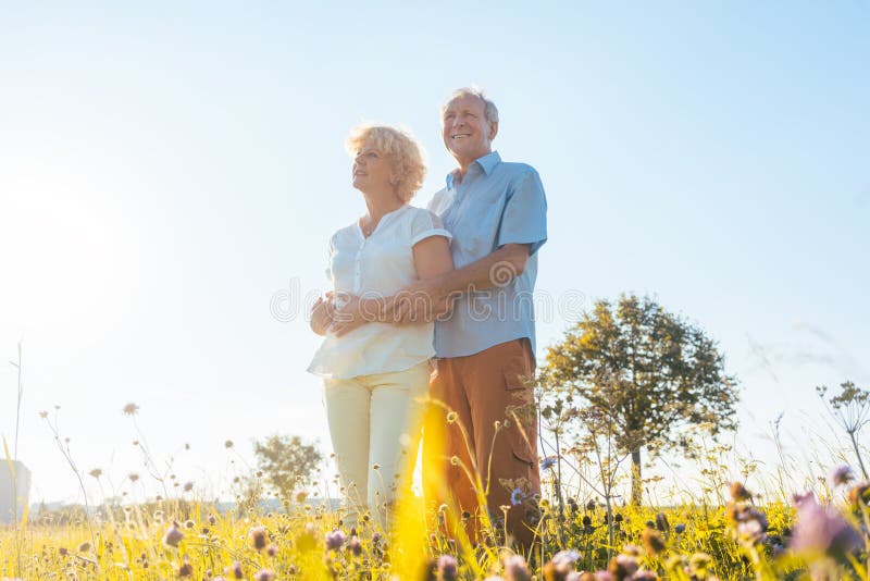 Low-angle view of a romantic elderly couple enjoying health and nature while standing together on a field in a sunny day of summer. Low-angle view of a romantic elderly couple enjoying health and nature while standing together on a field in a sunny day of summer