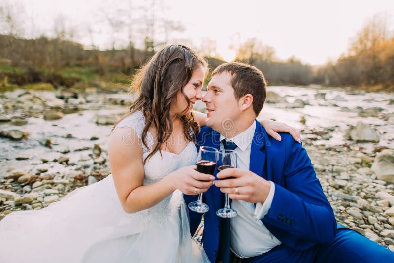 Romantyczna młoda bridal para pije wino na otoczaka brzeg rzeki z wzgórzami i strumienia jako tło zdjęcia royalty free Romantyczna młoda bridal para pije wino na otoczaka brzeg rzeki z wzgórzami i strumienia jako tło zdjęcia royalty free
