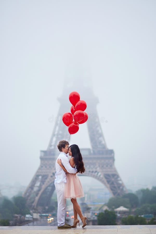 Beautiful romantic couple in love with bunch of red balloons together near the Eiffel tower in Paris on a cloudy and foggy rainy day. Beautiful romantic couple in love with bunch of red balloons together near the Eiffel tower in Paris on a cloudy and foggy rainy day