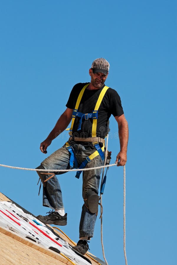 Roofer with safety harness shingling a roof with a steep pitch. Roofer with safety harness shingling a roof with a steep pitch.