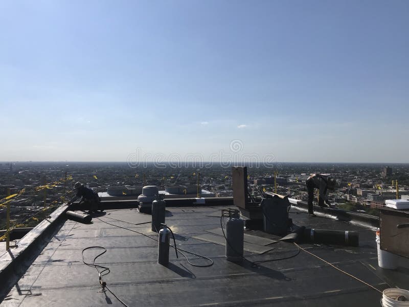 Roofer και ένα πλήρωμα που λειτουργεί σε μια ομαλή τροποποιημένη επίπεδη στέγη, πρόγραμμα υλικού κατασκευής σκεπής στοκ εικόνα Roofer και ένα πλήρωμα που λειτουργεί σε μια ομαλή τροποποιημένη επίπεδη στέγη, πρόγραμμα υλικού κατασκευής σκεπής στοκ εικόνα