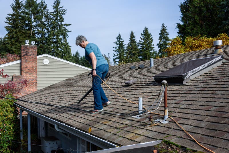 Rooftop view of suburban home, mature woman with leaf blower cleaning roof and gutter