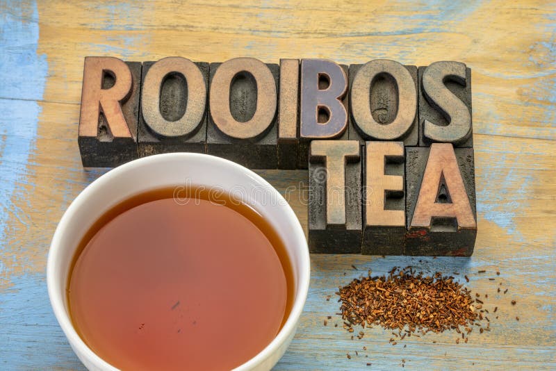 Rooibos red tea  -  a white cup of a hot drink, loose leaves and typography in letterpress wood type on grunge wood background, tea made from the South African red bush, naturally caffeine free. Rooibos red tea  -  a white cup of a hot drink, loose leaves and typography in letterpress wood type on grunge wood background, tea made from the South African red bush, naturally caffeine free